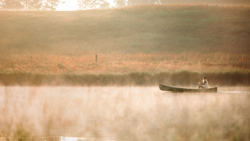 The image depicts a serene scene of a person paddling a canoe across a calm, mist-covered lake. The individual is positioned towards the right side of the image, seated in a small canoe with a single paddle in hand. The water reflects a soft glow from the warm, early morning or late afternoon light. In the background, a grassy hillside can be seen, with hues of green and brown gently blending into the misty atmosphere. The mist hovers just above the water’s surface