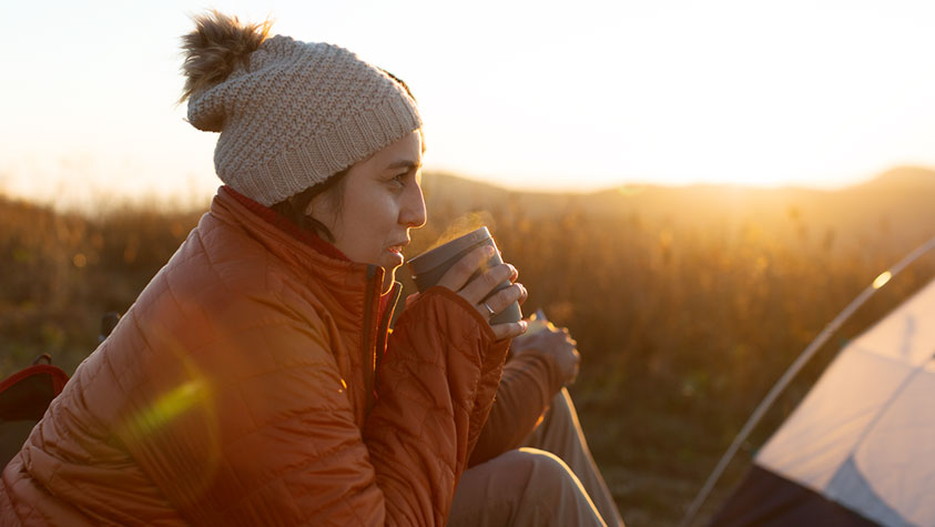 The image shows a person sitting outdoors at sunrise or sunset, sipping from a mug. They are wearing a knitted beanie with a pom-pom and a quilted orange jacket. The background includes tall grasses with a warm golden hue, indicating it is likely early morning or late afternoon. Part of a tent is visible to the right, suggesting a camping setting. The lighting casts a soft, golden glow across the scene.