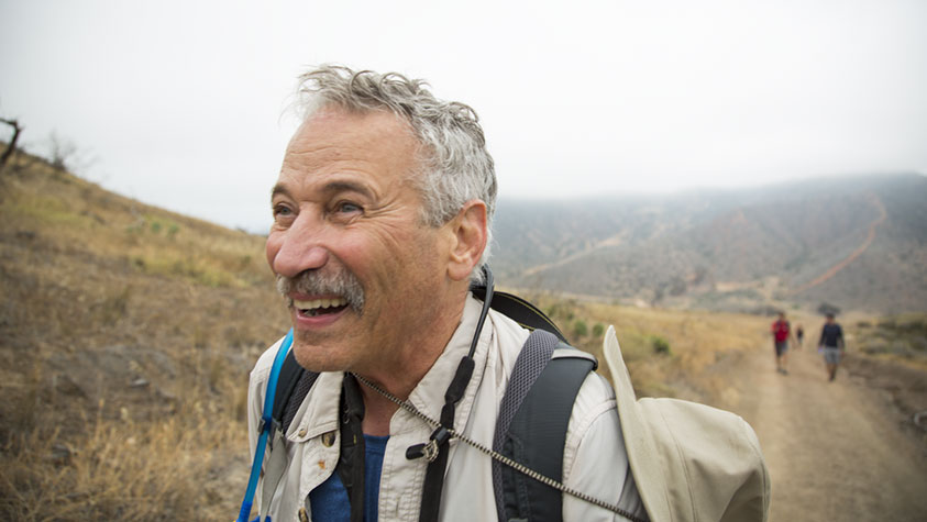 The image shows an older man with short gray hair and a mustache, walking along a dirt path in an arid, hilly landscape. He is smiling broadly, appearing joyful and energetic. He is wearing a light-colored jacket over a blue shirt, with a backpack and a pair of binoculars hanging around his neck. In the background, the terrain appears dry with patches of grass and sparse trees. The sky is overcast. Two more people, walking further behind on the trail, are blurred and less distinct.