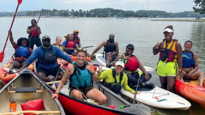 A diverse group of twelve people wearing swimsuits and swim shorts while sitting in boats with paddles in hand along the Tennessee RiverLine